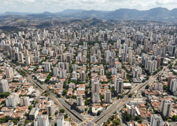 Do alto, Belo Horizonte ainda deixa aparecer a lógica traçada em prancheta no fim do século XIX. Ruas retas formam uma grade, avenidas cruzam o desenho em diagonais e a atual Avenida do Contorno acompanha o limite da zona urbana original. Entre o centro histórico e a Pampulha, a capital mineira transforma planejamento em paisagem cotidiana. Belo Horizonte nasceu de uma planta desenhada em 1895 A planta geral ficou pronta em 23 de março de 1895, quando o engenheiro Aarão Reis a apresentou ao governo mineiro. Segundo a Prefeitura de Belo Horizonte, a zona urbana tinha quarteirões de 120 m por 120 m. As ruas cruzavam-se em ângulos retos, enquanto algumas avenidas cortavam a malha em 45 graus. Porém, a famosa Avenida do Contorno não delimitava toda a área futura da capital. Ela separava a zona urbana planejada da zona suburbana, que seguia outra lógica de ocupação. O plano ainda previa uma zona rural além dessa faixa, reservada a sítios e pequena lavoura. Assim, o desenho visto do alto preserva sobretudo a estrutura central concebida pela Comissão Construtora da Nova Capital no fim do século XIX. 🏛️ 1894: A Comissão Construtora inicia os trabalhos da nova capital.🌿 1895: Aarão Reis conclui a planta geral da cidade planejada.🚀 1897: A nova capital de Minas Gerais é inaugurada em 12 de dezembro. Por que as diagonais cortam o tabuleiro de Belo Horizonte? As diagonais faziam parte de uma solução geométrica deliberada, não de um crescimento improvisado. Aarão Reis combinou ruas ortogonais com avenidas inclinadas, criando cruzamentos que quebravam a regularidade dos quarteirões. Portanto, a vista aérea revela duas ordens sobrepostas na mesma área central. Essa combinação continua fácil de reconhecer quando a malha regular encontra as grandes avenidas. O plano também diferenciava larguras. As ruas receberam 20 m e as avenidas 35 m. Uma grande avenida norte-sul recebeu 50 m. Aarão Reis ainda distribuiu nomes ligados a cidades, rios, montanhas e datas históricas de Minas Gerais e do Brasil. Hoje, caminhar pela região central ajuda a perceber essa lógica nos encontros entre vias retas e diagonais. A geometria não aparece apenas no mapa, ela interfere na orientação, nos cruzamentos e na sequência das quadras. Como enxergar o traçado original durante um passeio? O melhor caminho combina pontos dentro da Contorno com áreas que mostram outras fases urbanísticas da capital. Assim, o visitante percebe a cidade planejada sem transformar o roteiro em uma simples sequência de monumentos. O Parque Municipal nasceu ainda no processo de construção da nova capital, enquanto a Pampulha pertence a outra etapa urbana. Começar no centro e terminar na lagoa evidencia como Belo Horizonte incorporou novas ideias ao longo do século XX. Avenida Afonso Pena: atravessa parte importante da malha original e ajuda a visualizar a diferença entre ruas menores e grandes avenidas do plano. Praça da Liberdade: nasceu ligada ao centro administrativo e conserva a relação entre paisagismo, edifícios públicos e o desenho da nova capital. Parque Municipal Américo Renné Giannetti: inaugurado em 1897, oferece um contraste entre caminhos internos mais livres e a geometria rígida das ruas ao redor. Avenida do Contorno: funciona como referência prática para entender onde terminava a zona urbana prevista no projeto de 1895. Conjunto Moderno da Pampulha: mostra uma etapa posterior, criada em torno da lagoa e inscrita como Patrimônio Mundial em 2016. Quando ir a Belo Horizonte? O inverno costuma reunir os meses mais secos, enquanto o verão concentra os maiores volumes de chuva. Por isso, caminhadas pelo centro pedem atenção ao sol no período seco e à possibilidade de pancadas nos meses mais úmidos. .ant-grid { --cor-primaria: #006E00; /* Verde escuro */ --cor-secundaria: #000000; /* Preto para os destaques */ --cor-txt-secundaria: #B3B3B3; /* Cinza claro para os textos secundários */ display: grid !important; grid-template-columns: repeat(auto-fit, minmax(220px, 1fr)) !important; gap: 20px !important; justify-content: center !important; align-items: stretch !important; padding: 10px !important; margin: 30px auto !important; } .ant-card { border: 2px solid #1a3c1a !important; border-top: 5px solid var(--cor-txt-secundaria) !important; border-radius: 6px !important; background-color: var(--cor-primaria) !important; background-clip: padding-box !important; display: flex !important; flex-direction: column !important; overflow: hidden !important; cursor: pointer !important; box-shadow: 0 6px 16px rgba(0,0,0, 0.2) !important; transition: transform 0.2s ease, box-shadow 0.2s ease, border-color 0.2s ease !important; animation: wmFadeIn 0.6s cubic-bezier(0.2, 0.8, 0.2, 1) backwards; height: auto !important; min-height: 0 !important; margin: 0 !important; padding: 0 !important; } @keyframes wmFadeIn { from { opacity: 0; transform: translateY(30px); } to { opacity: 1; transform: translateY(0); } } .ant-card:hover { transform: translateY(-5px) !important; box-shadow: 0 12px 24px rgba(0, 110, 0, 0.3) !important; border-color: var(--cor-txt-secundaria) !important; } .ant-header { background: #004d00 !important; padding: 18px 10px !important; text-align: center !important; display: flex !important; flex-direction: column !important; align-items: center !important; justify-content: center !important; margin: 0 !important; border-bottom: 1px solid #1a3c1a !important; } .ant-body { background: #ffffff !important; padding: 18px !important; display: flex !important; flex-direction: column !important; flex-grow: 1 !important; align-items: center !important; margin: 0 !important; height: 100% !important; } .ant-card-destaque { border: 2px solid var(--cor-secundaria) !important; border-top: 5px solid var(--cor-secundaria) !important; box-shadow: 0 8px 20px rgba(0, 0, 0, 0.3) !important; } .ant-card-destaque .ant-header { background: var(--cor-secundaria) !important; } .ant-card-destaque .ant-header .ant-titulo { color: #ffffff !important; } .ant-badge-chuva { display: inline-flex !important; align-items: center !important; gap: 5px !important; background: #f0f5f0 !important; color: #555555 !important; font-size: 11px !important; font-weight: 700 !important; padding: 4px 10px !important; border-radius: 4px !important; margin-bottom: 12px !important; text-transform: uppercase !important; letter-spacing: 0.5px !important; border: 1px solid #dcdcdc !important; } .ant-badge-chuva.chuva-alta { background: rgba(204, 17, 17, 0.08) !important; color: #cc1111 !important; border-color: #ffcccc !important; } .ant-badge-chuva.tempo-seco { background: rgba(0, 110, 0, 0.08) !important; color: var(--cor-primaria) !important; border-color: #ccffcc !important; } .pill-ant { display: flex !important; justify-content: center !important; align-items: center !important; text-align: center !important; background: #e6f2e6 !important; color: var(--cor-primaria) !important; font-size: 11px !important; font-weight: 900 !important; letter-spacing: 0.5px !important; padding: 8px 16px !important; border-radius: 4px !important; text-transform: uppercase !important; margin-top: auto !important; margin-bottom: 0 !important; width: 100%; box-sizing: border-box; border: 1px solid #ccdccd !important;} .ant-card-destaque .pill-ant { background: var(--cor-secundaria) !important; color: #ffffff !important; font-weight: 900 !important; border: none !important; } .ant-emoji { font-size: 24px !important; line-height: 1 !important; margin-bottom: 6px !important; display: block !important; } .ant-titulo { color: #ffffff !important; font-size: 16px !important; text-transform: uppercase !important; margin-top: 5px !important; font-weight: bold !important; display: block !important; letter-spacing: 0.5px !important; } .ant-mes { font-size: 11px !important; font-weight: 700 !important; color: var(--cor-primaria) !important; text-transform: uppercase !important; margin-bottom: 8px !important; display: block !important; } .ant-card-destaque .ant-mes { color: var(--cor-txt-secundaria) !important; } .ant-temp { font-size: 20px !important; font-weight: 900 !important; color: var(--cor-primaria) !important; margin-bottom: 8px !important; display: block !important; text-align: center !important; } .ant-card-destaque .ant-temp { color: var(--cor-secundaria) !important; } .ant-desc { font-size: 12px !important; color: #444444 !important; text-align: center !important; line-height: 1.6 !important; margin-bottom: 15px !important; flex-grow: 1 !important; font-family: 'Segoe UI', Roboto, Helvetica, Arial, sans-serif !important; display: block !important;} .ant-desc strong { color: var(--cor-primaria) !important; font-weight: bold !important; } .ant-card-destaque .ant-desc strong { color: #000000 !important; } ☔ Verão Dezembro a Fevereiro 18°C a 27°C 🌧️ 124 a 268 mm/mês O que fazer: Intercalar áreas abertas com museus e espaços cobertos. Atenção: Concentra os maiores volumes de chuva, com frequentes pancadas que exigem um roteiro urbano mais flexível. Status: Estação chuvosa e úmida. ☔ MUSEUS E COBERTOS 🍂 Outono Março a Maio 13°C a 26°C 🌤️ 33 a 149 mm/mês O que fazer: Caminhar pelo centro e pela Praça da Liberdade. Vantagem: O clima começa a estabilizar e as chuvas diminuem significativamente, tornando os passeios muito mais confortáveis. Status: Transição para a época seca. 🍂 CENTRO E PRAÇAS 🌟 Inverno Junho a Agosto 11°C a 24°C ☀️ 6 a 12 mm/mês O que fazer: Explorar trajetos a pé e mirantes urbanos. Dica: É a época mais seca do ano, com sol firme e noites frias. Perfeito para caminhar pela cidade sem medo da chuva. Status: 🌟 Melhor Época! Clima firme e céu limpo. 🌟 TRAJETOS E MIRANTES 🌸 Primavera Setembro a Novembro 15°C a 27°C 🌤️ 49 a 195 mm/mês O que fazer: Combinar passeios pelo centro, parques e o complexo da Pampulha. Atenção: As temperaturas voltam a subir com força, acompanhadas pelo retorno progressivo das pancadas de chuva. Status: Aumento do calor e da instabilidade. 🌸 PARQUES E PAMPULHA As faixas usam mínimas e máximas climatológicas mensais e precipitação média. Consulte a página real do Climatempo antes da viagem. Um desenho que ainda organiza o olhar Belo Horizonte cresceu muito além da malha imaginada no século XIX, mas o centro ainda revela a disciplina geométrica do projeto original. Entre diagonais, quarteirões regulares e a Contorno, a história urbana permanece visível no cotidiano. Essa leitura ganha outra dimensão quando o visitante compara o centro planejado com a paisagem moderna da Pampulha e percebe duas épocas urbanísticas. Na próxima visita, escolha um ponto alto ou caminhe pelo centro e tente reconhecer esse tabuleiro por conta própria, mesmo sem mapa. Leia mais: Steve Jobs, “Seu tempo é limitado”, por que perceber que a vida acaba pode mudar a maneira como escolhemos vivê-la Por que cidades cada vez mais quentes estão clareando os telhados das casas, e o efeito não termina dentro dos imóveis Psicólogos investigam por que algumas pessoas estão cansadas, querem dormir, mas continuam adiando a hora de ir para a cama, estudos recentes tratam a chamada procrastinação do sono como problema de autorregulação
