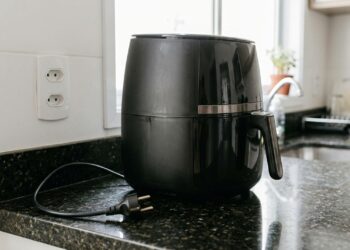 Airfryer preta desligada sobre bancada de cozinha, com plugue de três pinos desconectado de tomada na parede, ilustrando uma medida de segurança elétrica residencial. - (Imagem gerada por inteligência artificial-ChatGPT/Olhar Digital)