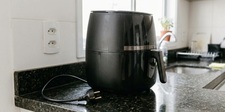 Airfryer preta desligada sobre bancada de cozinha, com plugue de três pinos desconectado de tomada na parede, ilustrando uma medida de segurança elétrica residencial. - (Imagem gerada por inteligência artificial-ChatGPT/Olhar Digital)