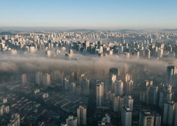 Vista aérea de São Paulo com prédios parcialmente encobertos por uma camada espessa de poluição acinzentada durante uma manhã de inverno, ilustrando os efeitos da inversão térmica. - (Imagem gerada por inteligência artificial-ChatGPT/Olhar Digital)