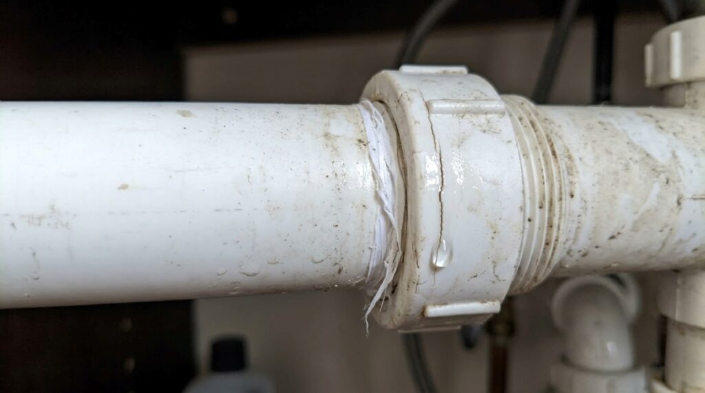 Conexão hidráulica de PVC branco com pequena rachadura próxima à rosca e gota de água vazando, mostrando um possível dano causado por aperto excessivo. - (Imagem gerada por inteligência artificial-ChatGPT/Olhar Digital)