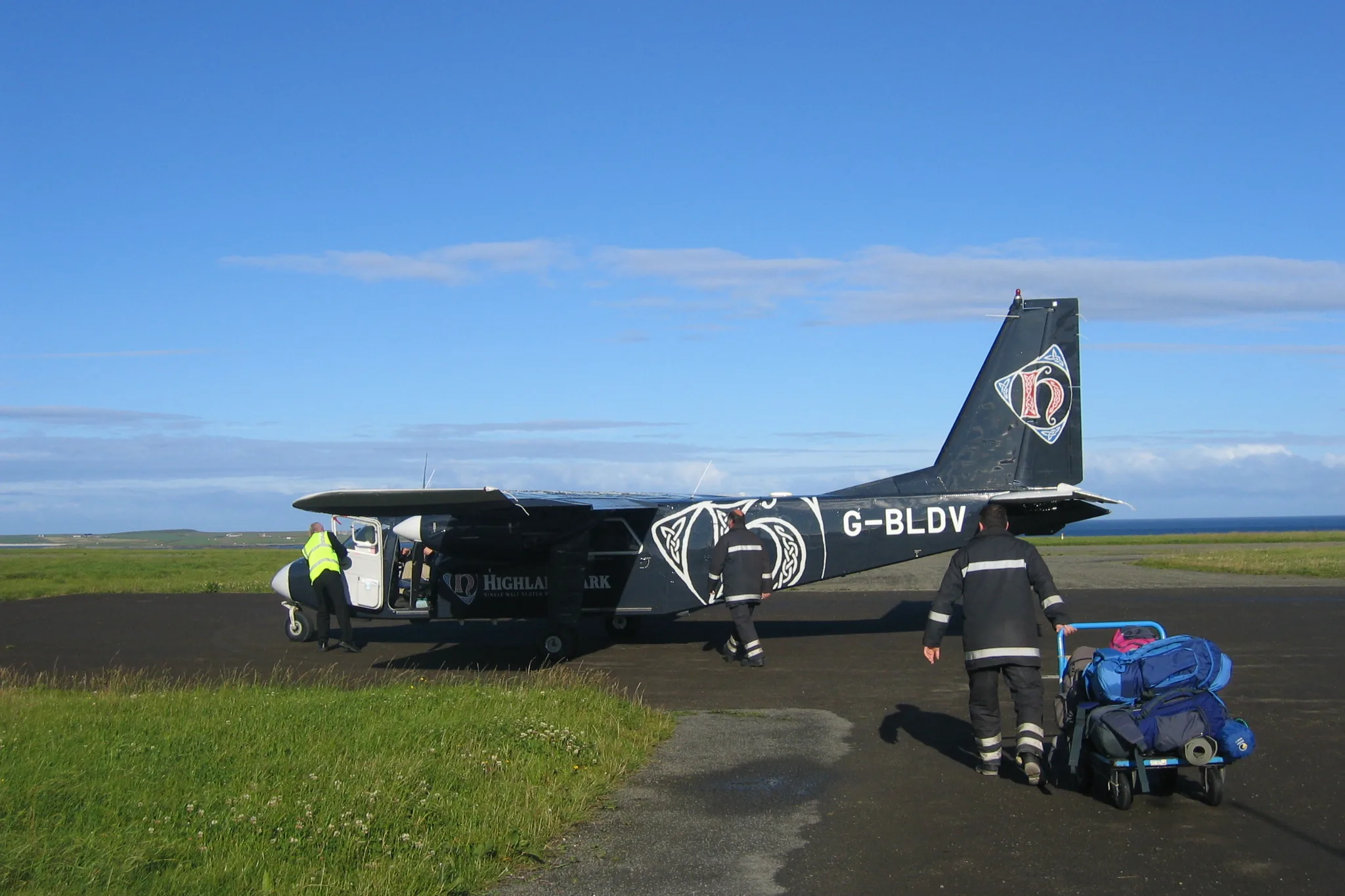Foto de um Islander estacionado no pequeno aeródromo de Papa Westray, com a porta aberta, um funcionário ao lado e outro empurrando um carrinho de bagagem pela pista de cascalho.