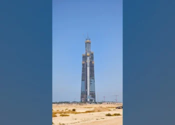 Foto da Jeddah Tower em construção em maio de 2026: torre esbelta e afilada no deserto, com vidro nos andares inferiores, núcleo de concreto e três gruas no topo, apresentada sobre fundo desfocado da própria imagem.