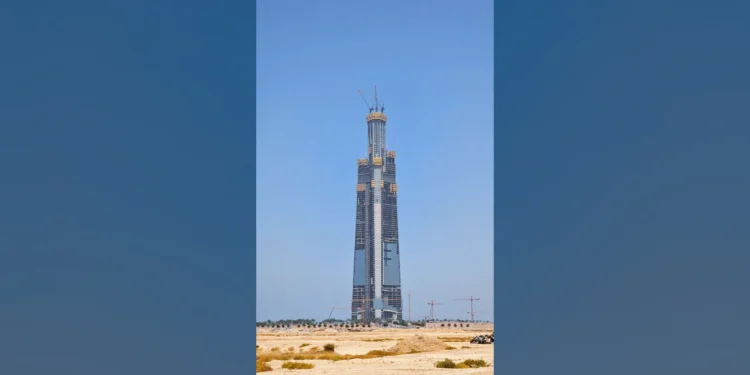 Foto da Jeddah Tower em construção em maio de 2026: torre esbelta e afilada no deserto, com vidro nos andares inferiores, núcleo de concreto e três gruas no topo, apresentada sobre fundo desfocado da própria imagem.