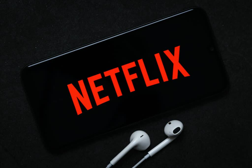 Logo da Netflix é exibida em tela de smartphone, com fones de ouvido ao lado do aparelho