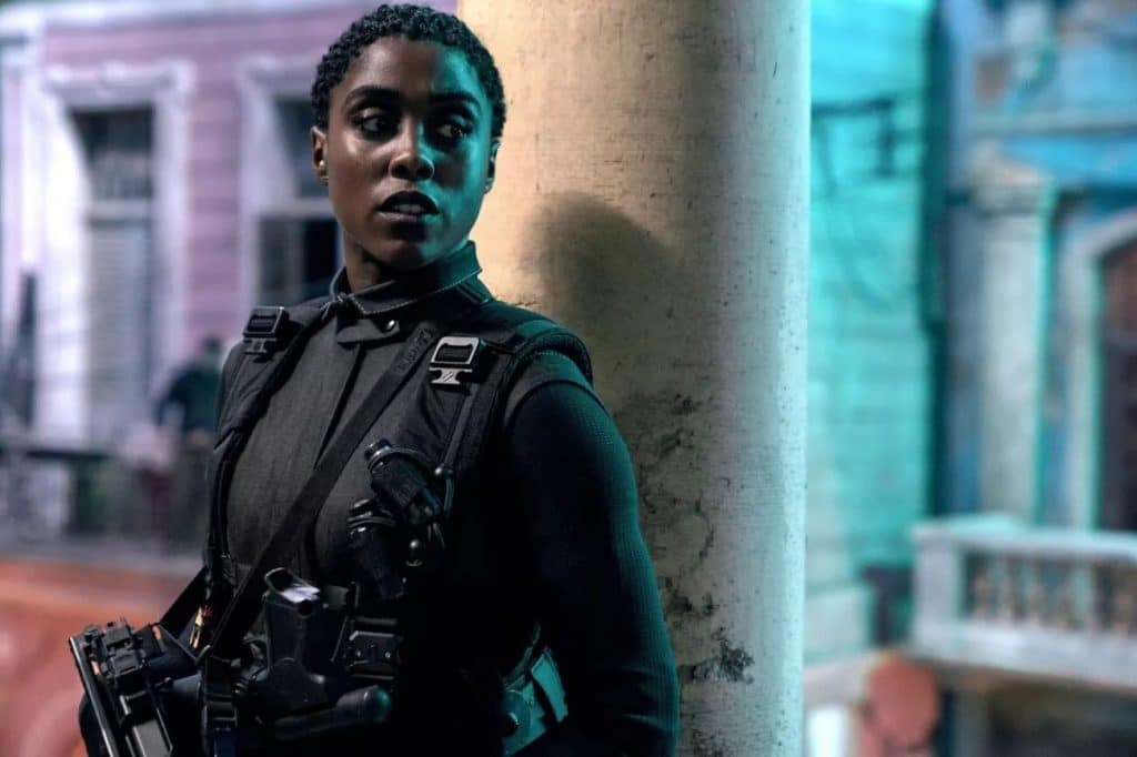 Imagem mostra a atriz Lashana Lynch, do filme "007: No Time to Die", vestida com roupa de infiltração militar e armada com uma submetralhadora, encostada em uma pilastra cinza