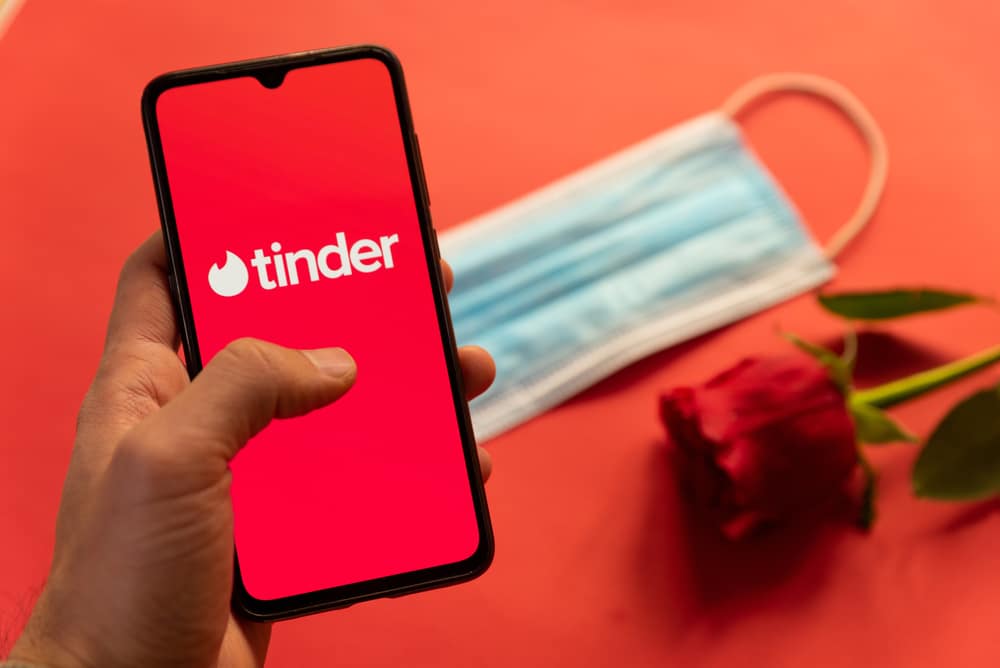 Celular com o símbolo do Tinder na tela e ao fundo uma máscara de proteção facial e uma flor
