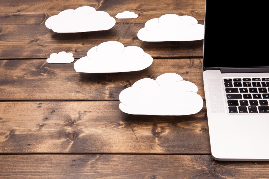 Imagem mostra uma parte de um notebook, em cima de uma superfície de madeira, e ao lado dele algumas nuvens feitas de papel. A ideia é ilustrar a computação em nuvem.