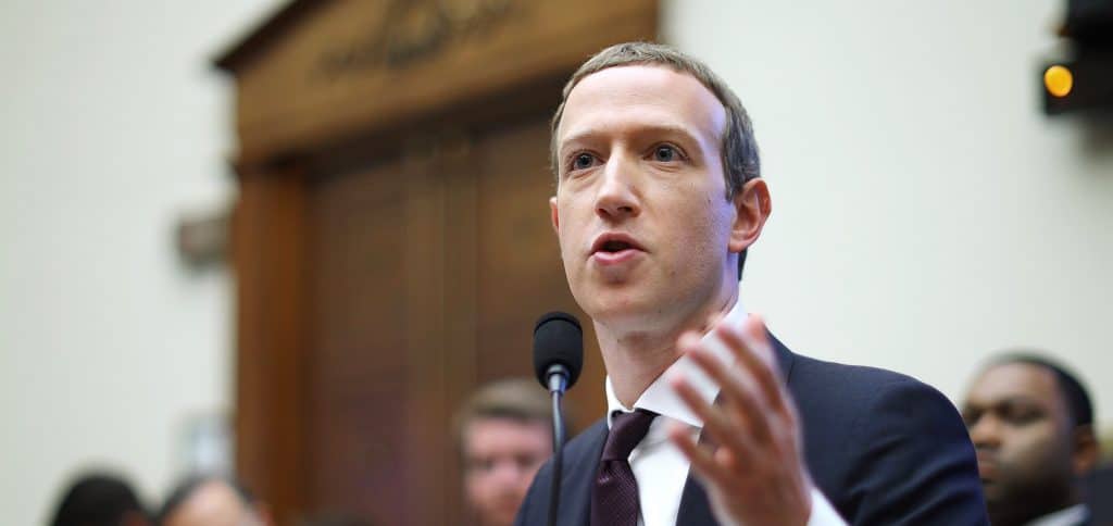 Inagem mostra o CEO do Facebook, Mark Zuckerberg, sentado em um acadeira de madeira, respondendo a perguntas durante uma audiência. Ele veste terno preto com gravata da mesma core está com o cabelo curto