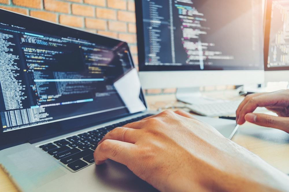 Imagem mostra uma mão digitando um código de programação em um teclado de notebook, ao lado a tela de um iMac.