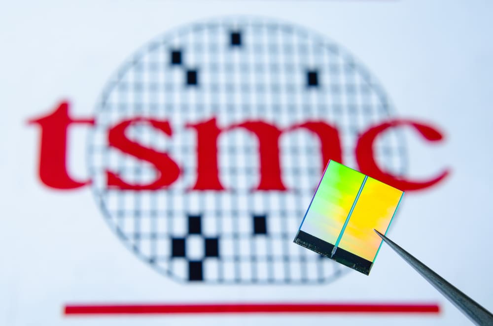 Imagem mostra ao fundo o logotipo da Fabricante de chips TSMC, à frente há uma pinça segurança um chip. Fornecedora da Apple planeja expansão no Arizona