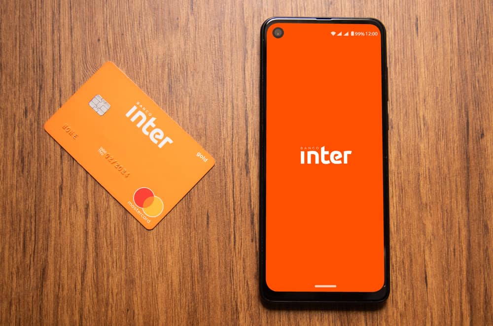 Em uma superfície de madeira, há um cartão do Banco Inter e, ao lado, um smartphone exibindo o logo do mesmo banco na tela.