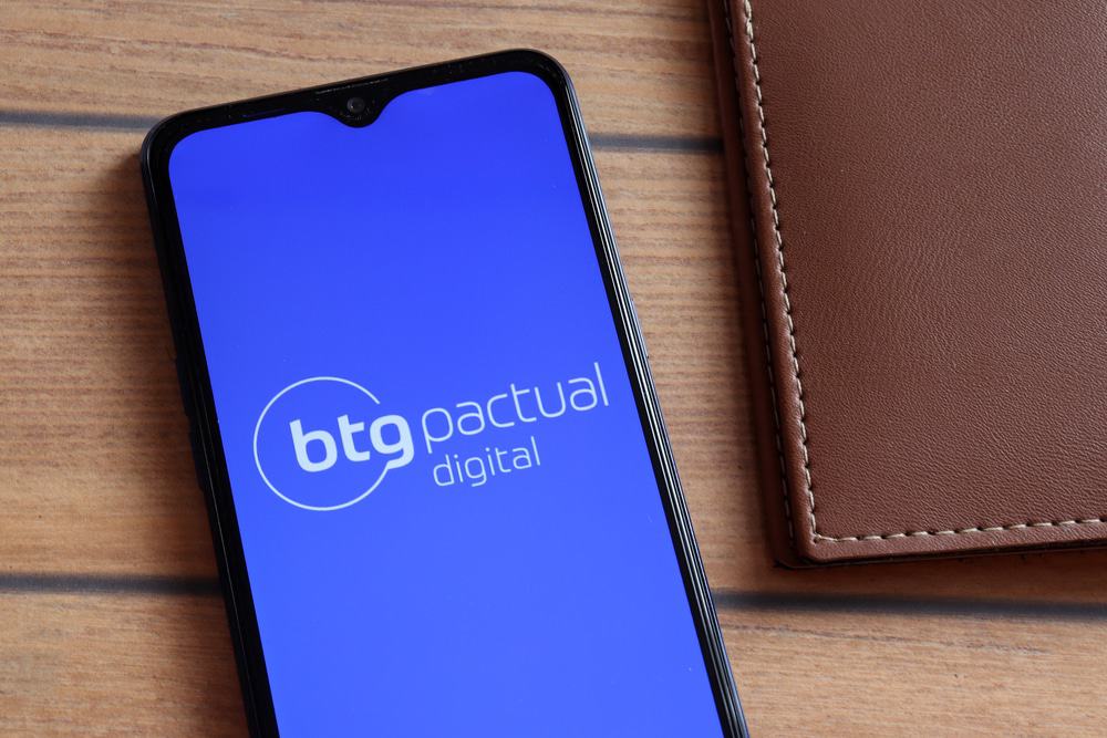 Imagem mostra um smartphone com o logo do BTG Pactual na tela, ao lado um pedaço de uma carteira de couro; ambos estão sobre uma superfície de madeira