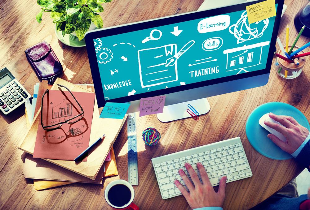 Imagem ilustra treinamento e capacitação em tecnologia, com uma mesa cheia de papeis e um monitor acompanhado de mouse e teclado.