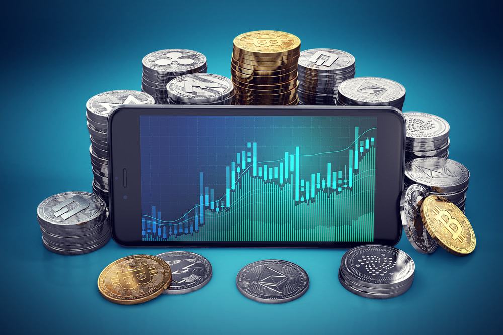 Imagem exibe um celular com a tela cheia de gráficos; ao fundo, pilhas de diferentes criptomoedas