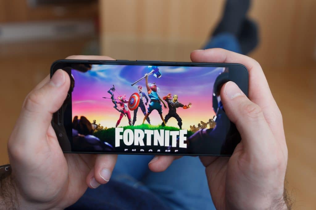 Imagem mostra um celular sendo segurado na horizontal por duas mãos masculinas. Na tela do aparelho, a logomarca do jogo Fortnite é exibida