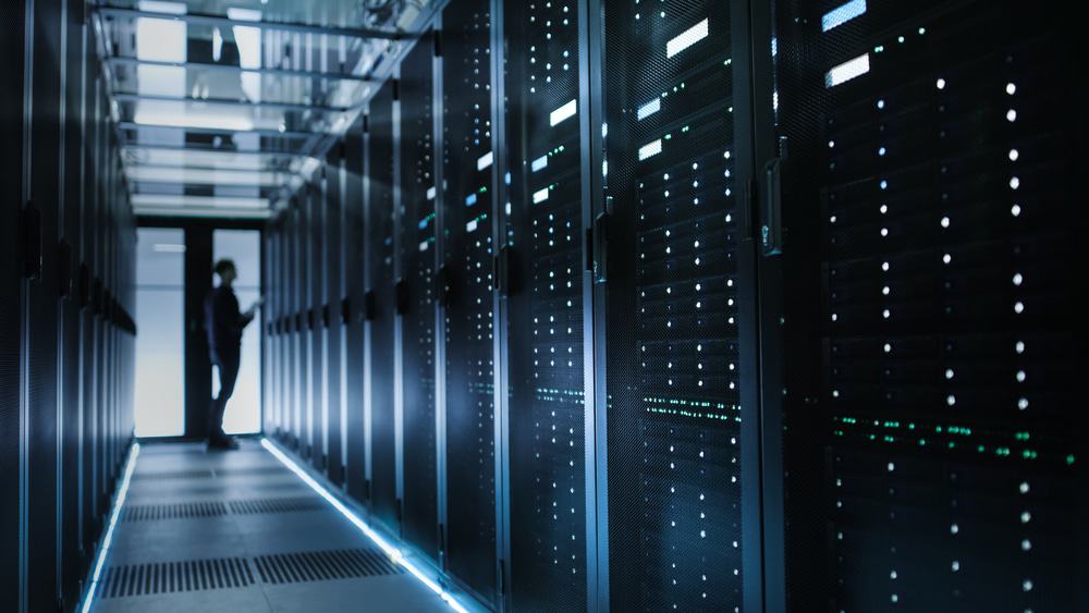 Foto do corredor de um data center; ao fundo, há a silhueta de um técnico de TI que está observando a infraestrutura tecnológica.