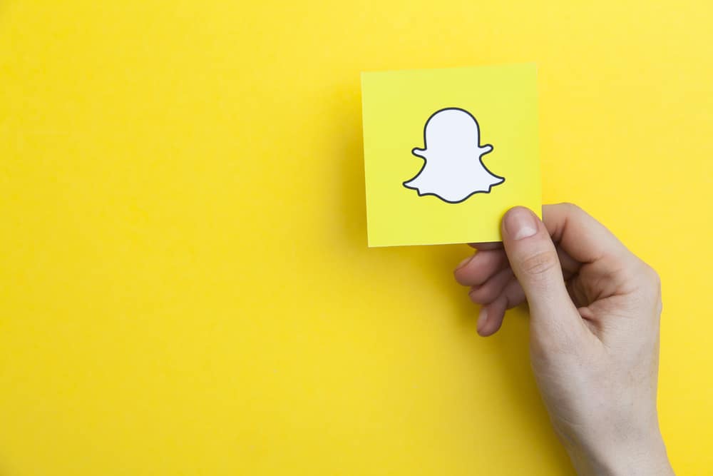 Em fundo amarelo, imagem mostra uma mão segurando um cartão que tem uma ilustração do fantasma, logomarca da rede social Snapchat