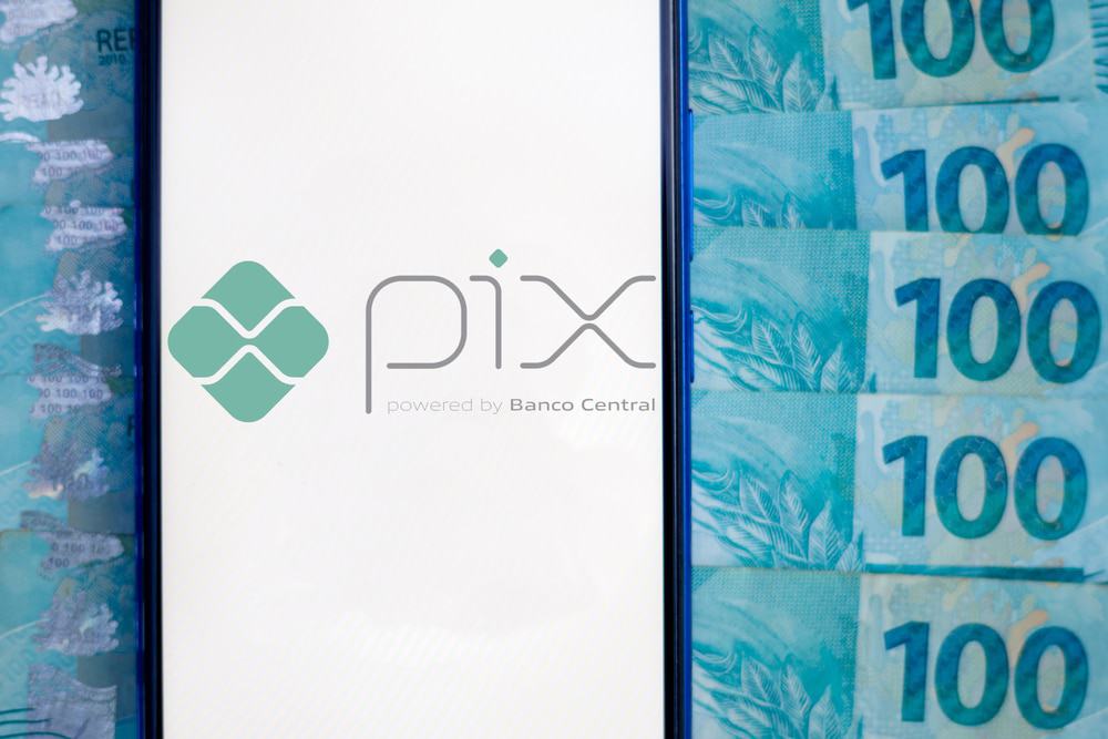 Imagem mostra a tela de um smartphone exibindo o logotipo do sistema de pagamentos instantâneo do Banco Central, o PIX; ao fundo aparecem várias notas de cem reais.
