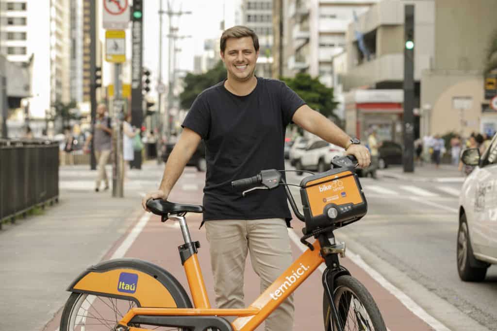 Foto mostra o fundador da Tembici, Tomás Martins, segurando uma das bikes da empresa em parceria com o Itaú; ao fundo está uma avenida