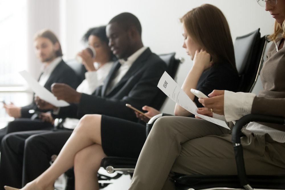 Cinco pessoas estão sentadas, uma ao lado das outras, esperando para serem chamadas para a entrevista de emprego.
