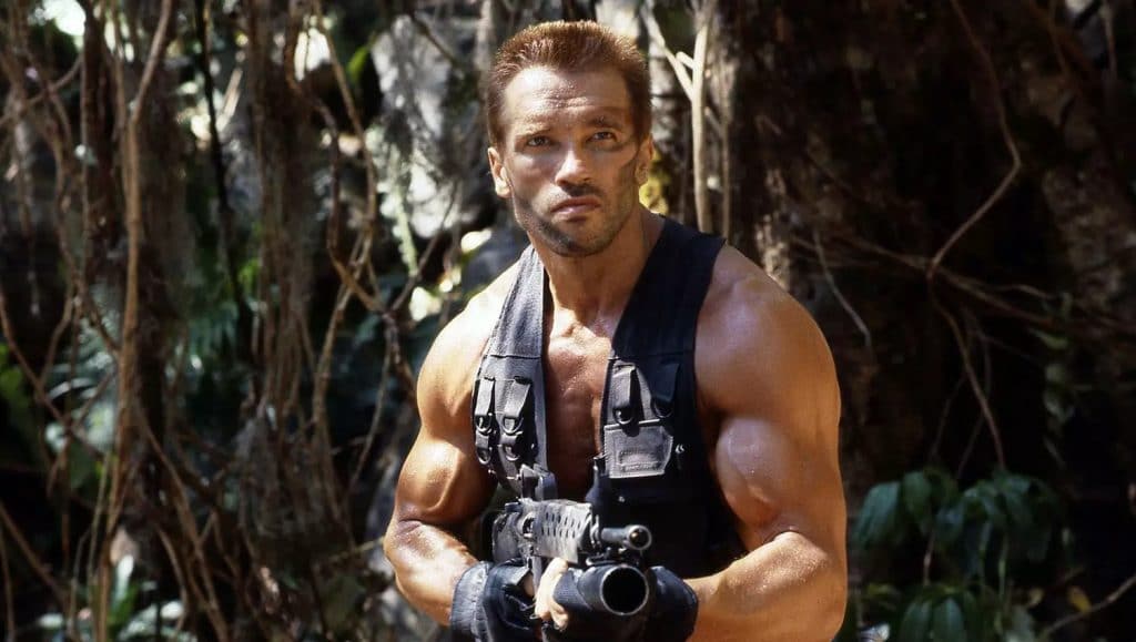 Foto mostra Arnold Schwarzenegger em cena do primeiro filme, em uma selva.