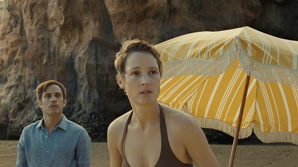Cena do filme mostra Guy (Gael García Bernal) e Prisca (Vicky Krieps) na praia com olhar assustado.