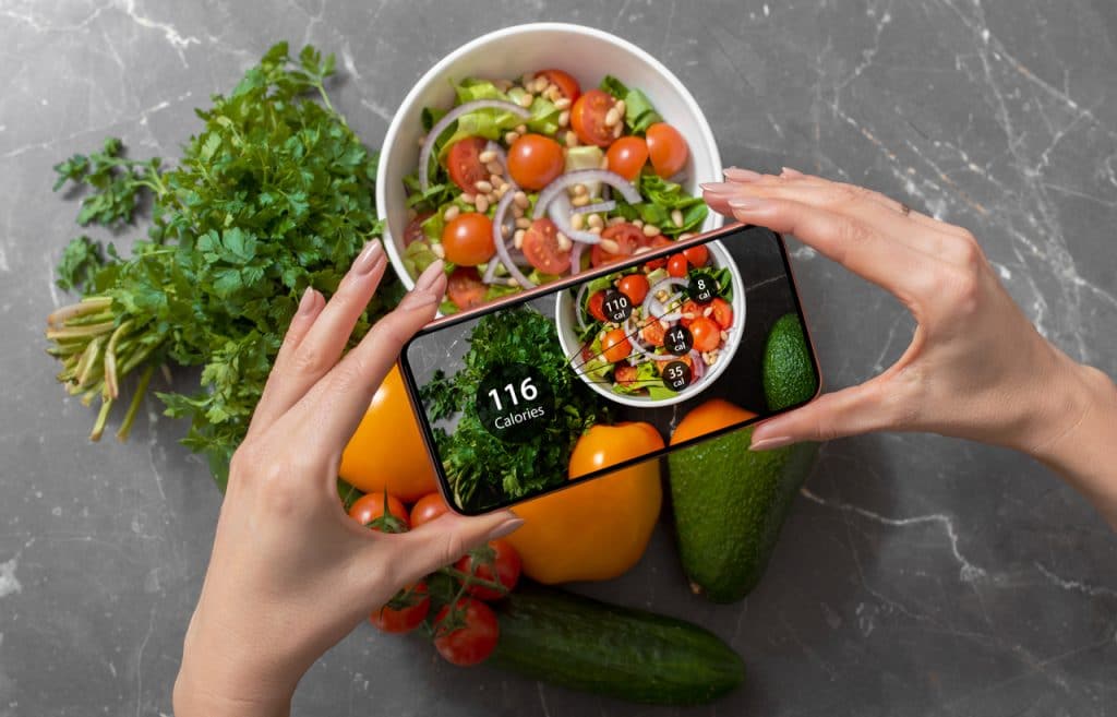 Imagem mostra um prato de salada sob um fundo cinza de pedra. À frente, duas mãos femininas seguram um smartphone, que está tirando foto da comida