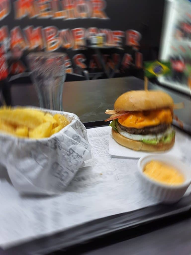 Foto mostra um hambúrguer artesanal, acompanhado de batatas e molho. A imagem tem um filtro de desfoque, gerado pela inteligência artificial do smartphone Galaxy M62