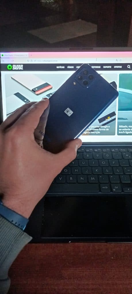 Imagem mostra a parte traseira do Galaxy M62, sendo segurado por uma mão e um notebook exibindo a página inicial do Olhar Digital ao fundo. Samsung lançou o Galaxy M32 e o Galaxy M62 no varejo online brasileiro em 2 de agosto
