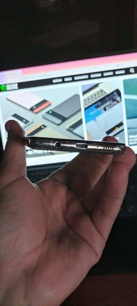 Imagem mostra a parte inferior e as entradas de fone e USB-C do Galaxy M62, sendo segurado por uma mão e um notebook exibindo a página inicial do Olhar Digital ao fundo. Samsung lançou o Galaxy M32 e o Galaxy M62 no varejo online brasileiro em 2 de agosto