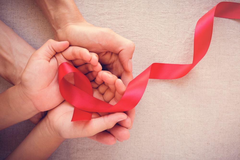 Imagem mostra quatro mãos, umas em cima das outras, com as palmas para cima também, segurando uma fita vermelha que é símbolo do combate ao vírus HIV causador da Aids