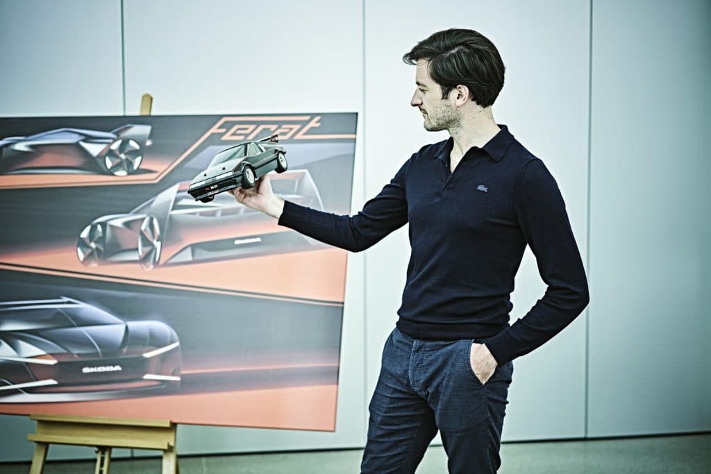 O designer Baptiste Brugiere segurando um mini modelo do Skoda Ferat ao lado de uma tela com desenhos do seu conceito do carro