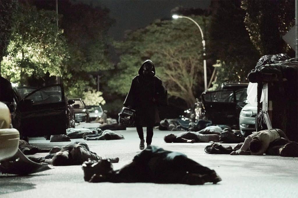 Foto mostra personagem utilizando capa e máscara de proteção biológica enquanto caminha por rua escura repleta de corpos pelo chão.