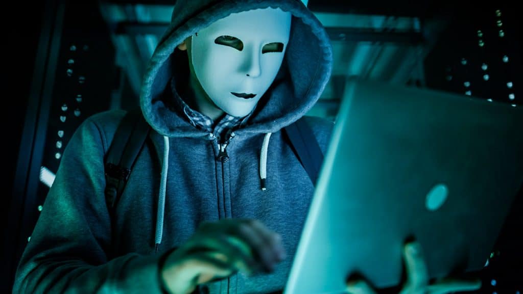 hacker com máscara e moletom de capuz digitando em notebook