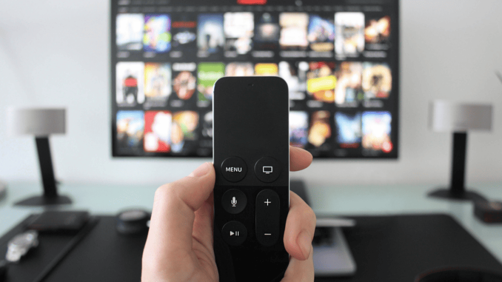 Imagem de uma mão segurando um controle remoto, com uma TV ao fundo exibindo vários programas