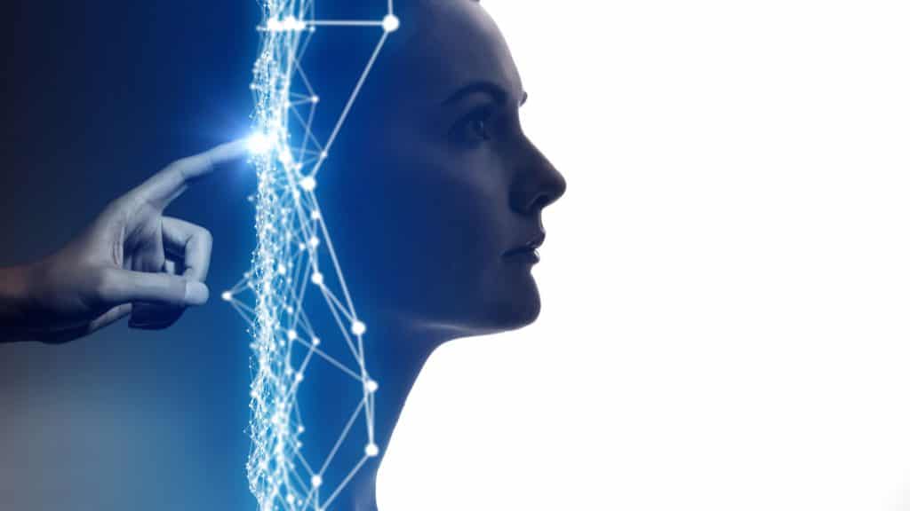 Imagem conceitual mostra uma mão tocando com o indicador a parte de trás da cabeça de uma mulher, com traços digitais azuis entre um e outro. A ideia é simbolizar a inteligência artificial