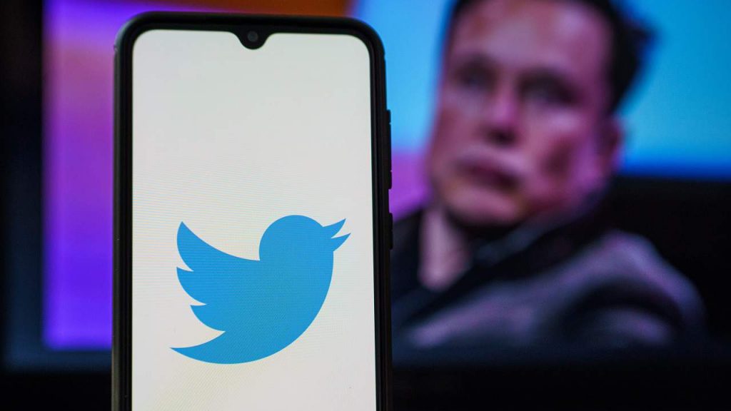 Celular com logomarca do Twitter aberta sobre fundo branco e, ao fundo, foto de Elon Musk aberta em tela de computador