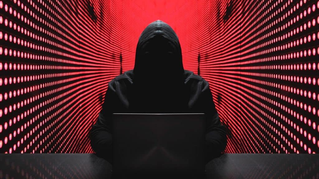 Hacker encapuzado sentado usando notebook numa mesa e, ao fundo, paredes com luzes vermelhas