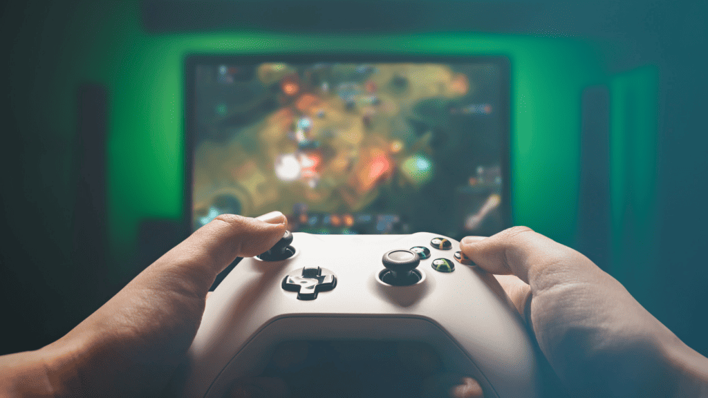 Mãos de um jogador desconhecido empunhando uma manete de videogame com uma tela ao fundo