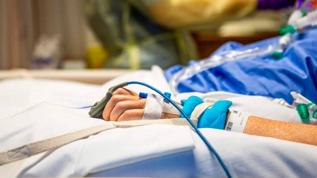 Braço de paciente estendido sobre as pernas numa cama de hospital