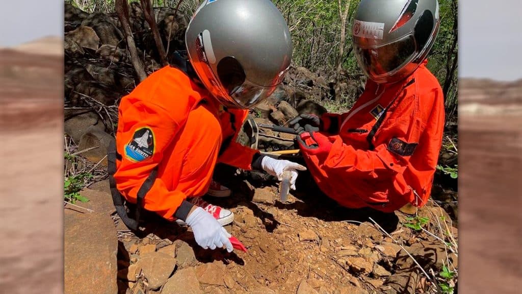 Duas pessoas com trajes de astronautas agachadas enquanto uma coleta amostras do chão no projeto Habitat Marte