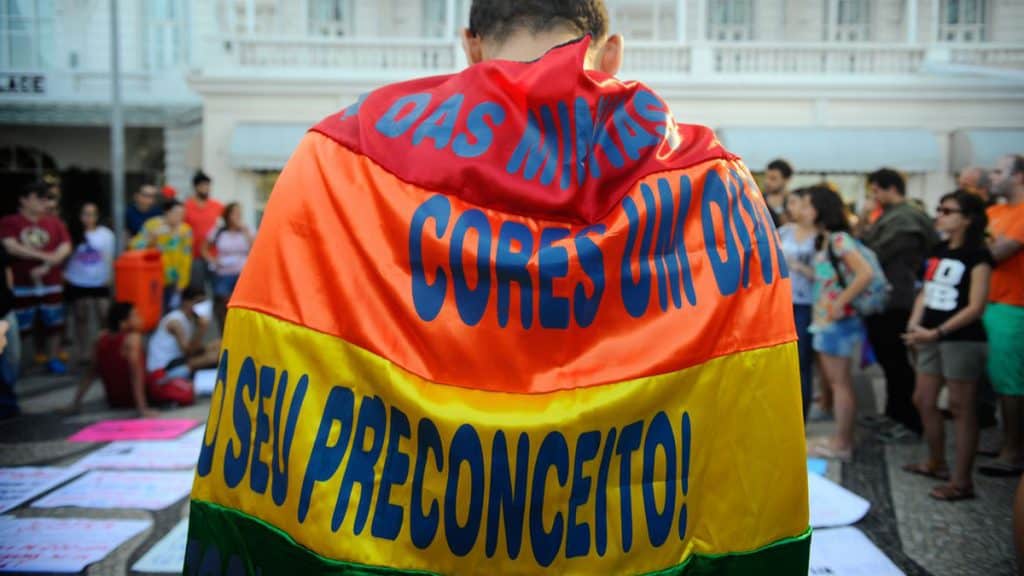 Homem de costas vestindo bandeira LGBTQIAP+ em parada na rua