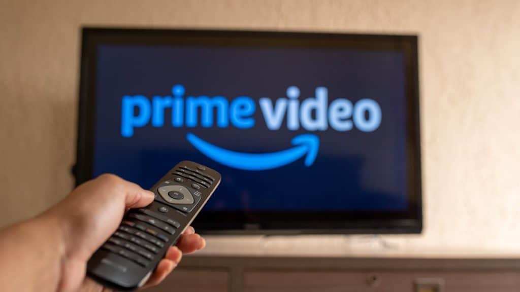 Pessoa segurando controle de TV e apontando para a TV, que exibe a logomarca do Prime Video, da Amazon, na tela