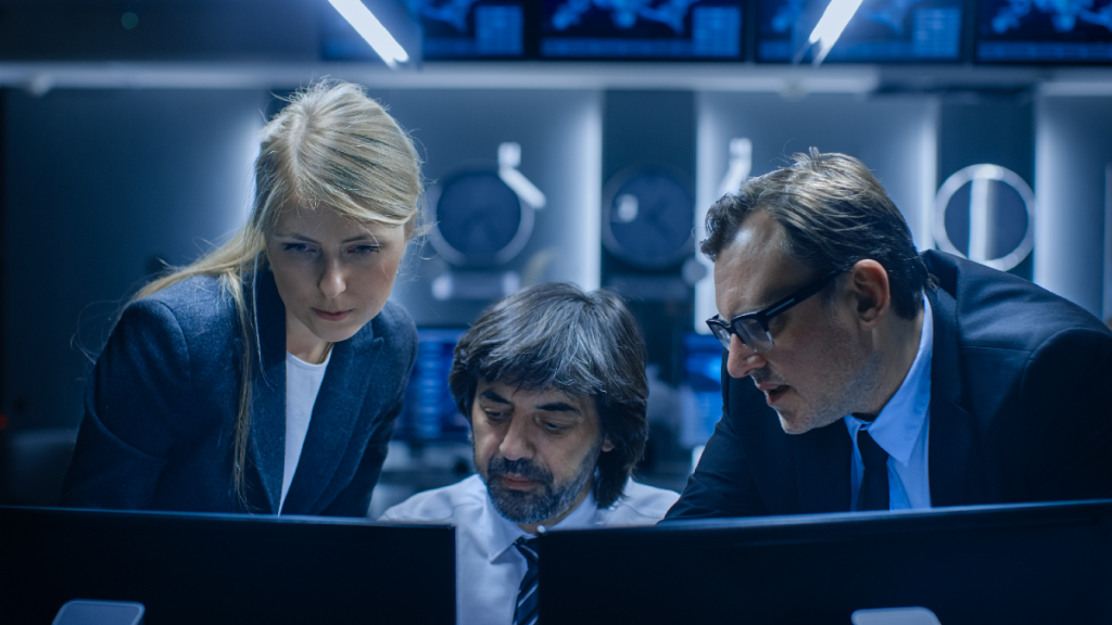 Uma mulher e dois homens encarando duas telas de computadores, á sua frente.