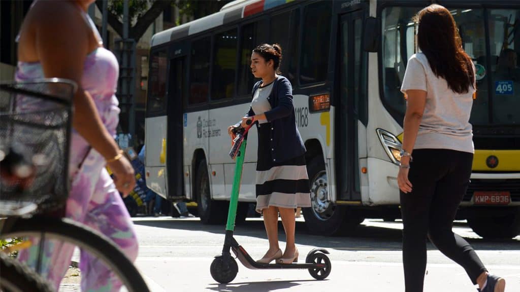 Mulher andando de patinete elétrico no centro da cidade com pessoas e ônibus ao redor