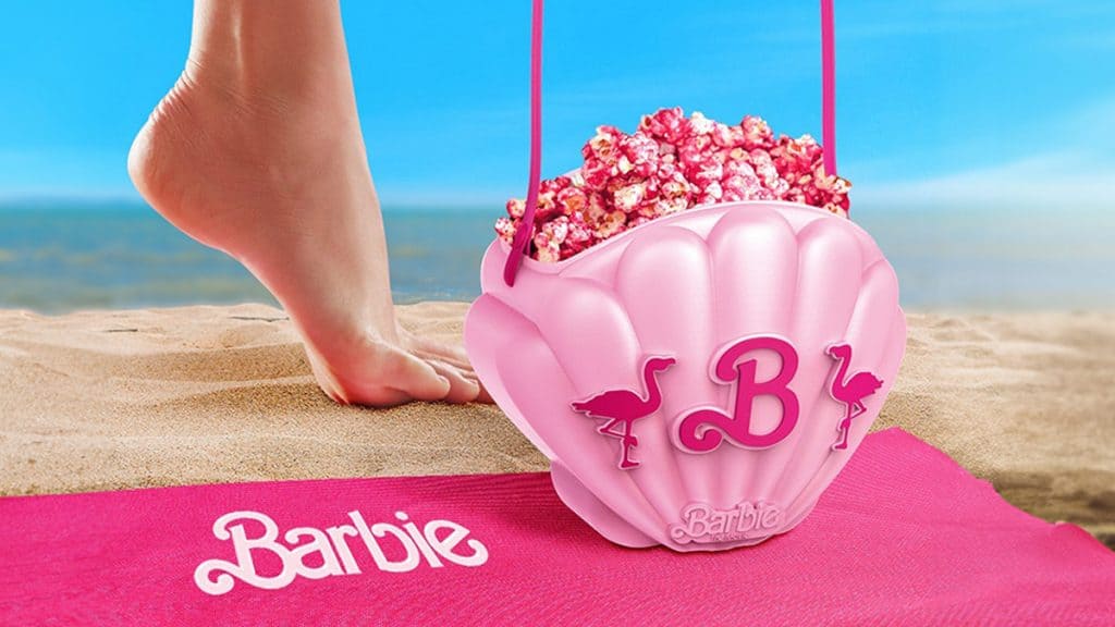 Imagem mostra balde de pipoca rosa em forma de concha com símbolo da Barbie, areia e um pé em destaque