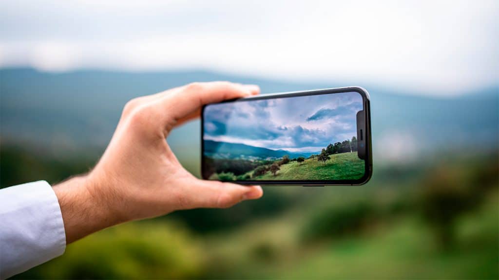 Tem um iPhone? Confira 4 funções secretas do botão de volume que muitos desconhecem Você sabia que os tradicionais botões de volume, presentes no lado esquerdo dos modelos de iPhone, possuem outras funções interessantes além de aumentar ou diminuir o som? Pois é, a seguir, vamos mostrar 4 funcionalidades secretas que são possíveis de realizar em seu aparelho utilizando apenas esses componentes. Pessoa tirando foto de paisagem com iPhone