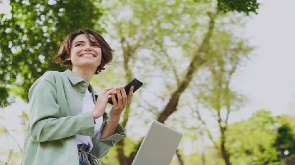 mulher com celular e notebook em meio a natureza sorrindo