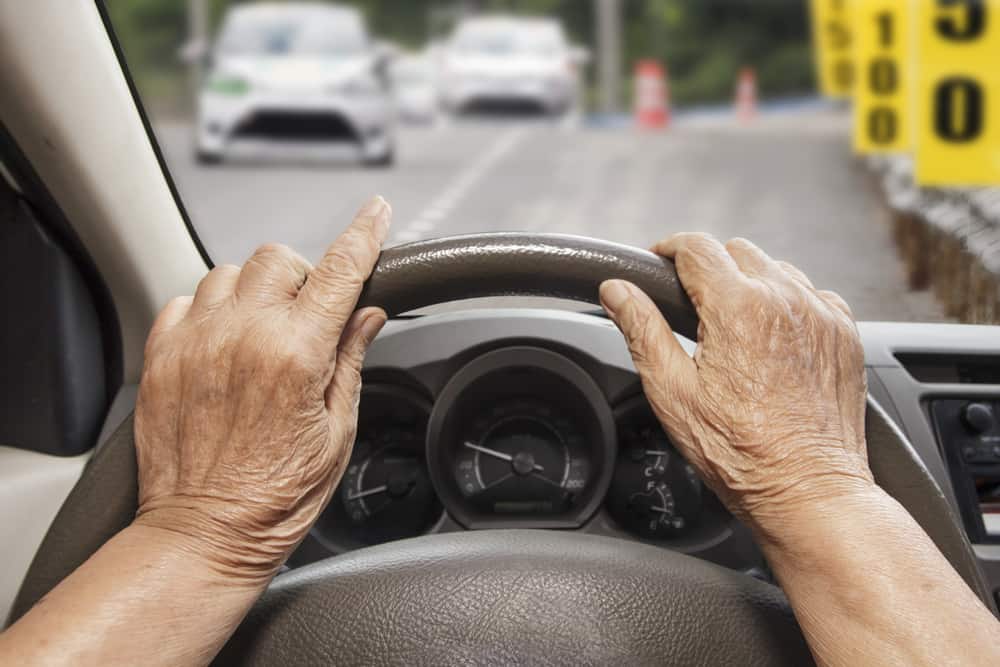 Mãos de uma pessoa segurando um volante, com imagem desfocada de uma rua com dois carros em segundo plano